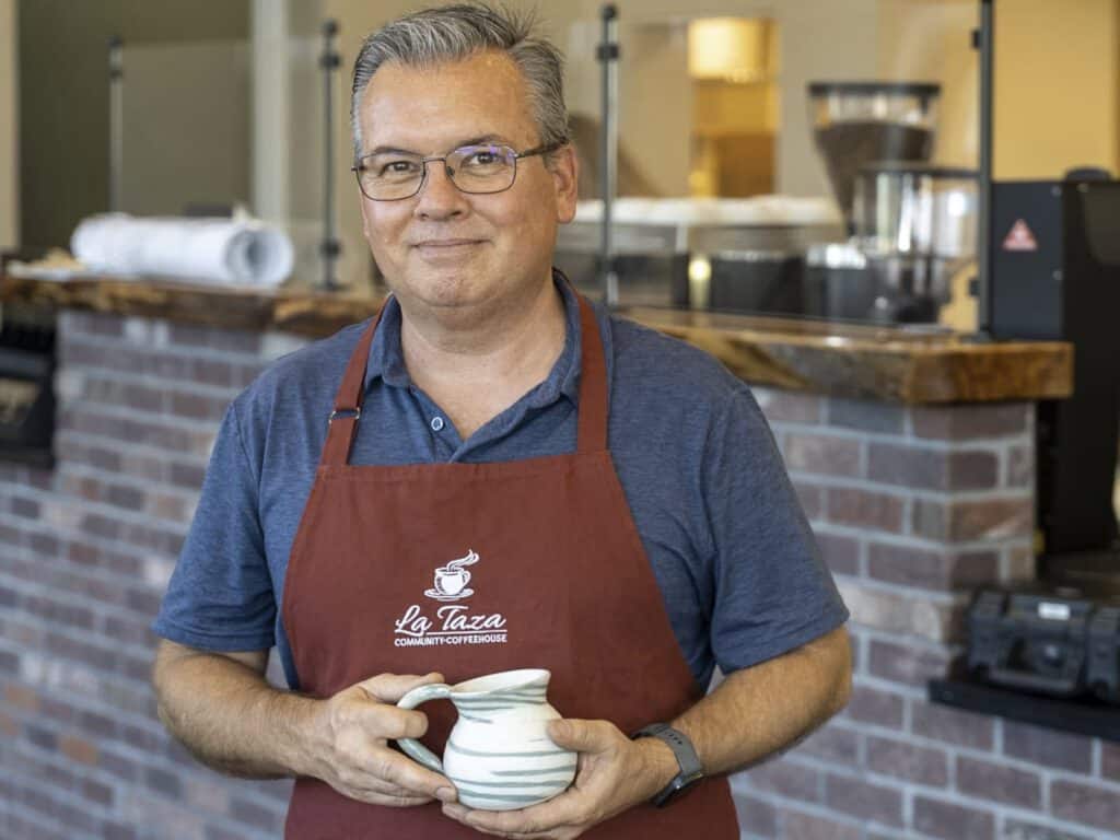 Man smiling with coffee mug at La Taza Community Coffeehouse launch event.